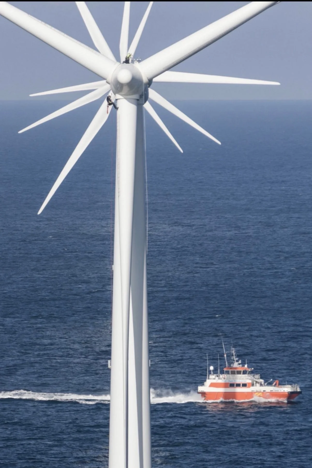 Technicians working on an offshore wind turbine with a support vessel below